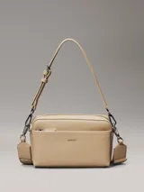 Cartera rectangular tipo bandolera de piel sintética color beige claro, con bolsillo exterior frontal y cierre superior de cremallera. Incluye dos correas desmontables: una ancha y una fina ajustable. Presenta el logo metálico de Calvin Klein en la parte frontal.