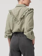 Blusa verde de algodón orgánico con cuello camisero, manga larga y cierre frontal de botones. Presenta volantes bordados que recorren el frente, la espalda y las sisas, además de detalles bordados en toda la prenda.