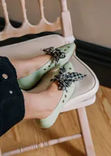 Bailarina de piel natural en color verde menta, con diseño de punta redondeada y detalle de ojales metálicos. Incluye un lazo desmontable de tela con estampado de cuadros vichy en blanco y negro.