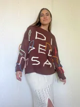 Sweater de punto color marrón con diseño oversize, cuello redondo y mangas largas. Presenta bordados artesanales en relieve con hilos de colores y detalles en lurex, incluyendo flecos decorativos distribuidos por toda la prenda.