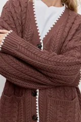 Cardigan de punto con diseño de trenzas, color marrón, con cierre frontal de botones y detalle de borde en color blanco. Presenta bolsillos frontales y mangas largas.
