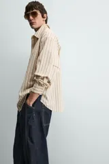 Camisa de algodón de corte relajado, color beige con finas rayas verticales en tonos gris y amarillo. Presenta cuello solapa, manga larga con puños abotonados y bajo con aberturas laterales.