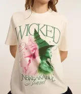 Remera color crema de algodón con cuello redondo y mangas cortas. Estampado frontal con imágenes del musical Wicked y texto en verde y rosa.