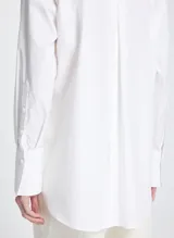 Camisa blanca de corte recto y estilo oversized, con cuello clásico, mangas largas con puños anchos, bolsillo delantero, cierre frontal con botones ocultos y bajo ligeramente redondeado.