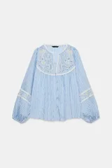 Blusa de rayas verticales blancas y celestes, con cuello redondo y escote en pico. Presenta detalles de bordados perforados y puntillas en el pecho y las mangas. Tiene mangas largas abullonadas con puños elásticos y cierre frontal con botones forrados.