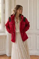 Piloto impermeable rojo con capucha integrada, cintura regulable con cordón y tranca, y mangas con volumen. Presenta detalles de volados estructurales en color verde en los hombros y puños elastizados.