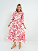 Vestido camisero midi de algodón con estampado floral en tonos rosados y blancos. Tiene cuello camisero, mangas 3/4, botones frontales, falda escalonada y cinturón fino.