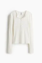 Remera ajustada de punto acanalado suave, color blanco, con cuello redondo ribeteado en picot y tapeta de botones. Es de manga larga.