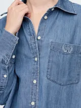 Camisa de jean de corte holgado, con cuello clásico, cierre frontal de botones y bolsillo en el pecho con logo bordado. Presenta mangas largas con puños anchos.