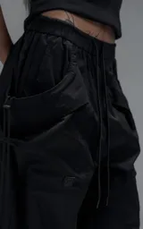 Pantalón de estilo utilitario y corte oversize, confeccionado en textil técnico liviano. Presenta cintura elástica con cordón ajustable, bolsillos laterales envolventes de gran tamaño con cordones y tancas de ajuste, y elásticos regulables en los tobillos.