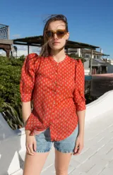 Camisa de estilo romántico color rojo con estampado floral pequeño en tono azulado, cuello redondo, cierre frontal con botones y mangas al codo con frunces en los hombros.