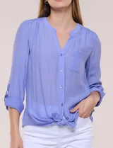 Blusa violeta de corte fluido, con cuello mao, escote en V y bolsillo plaqué en el pecho. Presenta mangas largas regulables con presilla y un lazo frontal para anudar en la cintura.
