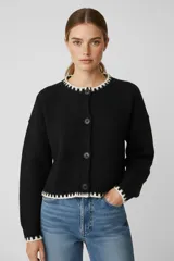 Saco de punto negro con diseño holgado, cuello redondo y cierre frontal con botones. Presenta detalles de contraste en color blanco en el cuello, puños y ruedo.