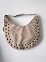 Cartera tipo hobo de símil gamuza color beige, con diseño de media luna y decorada con tachas metálicas plateadas en todo su contorno. Incluye correa ajustable y bolsillo interno.