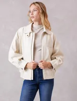 Campera bomber de eco cuero color beige, con cuello camisero, cierre frontal metálico y bolsillos delanteros con solapa y botón a presión. Presenta mangas largas con puños abotonados y ruedo trasero elastizado.