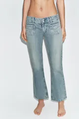 Jeans de tiro bajo con corte flare, confeccionados en denim azul con lavado localizado. Presentan bolsillos de plastrón en el frente, trabillas para cinturón y cierre frontal con cremallera y botones metálicos.