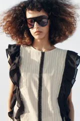 Blusa de cuello redondo y manga sisa, de color beige con rayas verticales. Presenta volantes negros en las sisas y una tira de encaje central con jaretas. Cuenta con cierre en la espalda con abertura y botón.