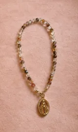 Collar compuesto por una cadena de piedras naturales facetadas en tonos translúcidos, beige y tierra, con un dije central de la Medalla Milagrosa en metal dorado.