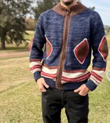 Campera de punto con cierre frontal, cuello alto y diseño artesanal. Presenta un cuerpo en tono azul jaspeado con detalles en marrón, blanco y rojo, incluyendo parches en forma de rombo en el pecho y los codos.