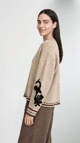 Sweater de lana color beige con calce holgado y cuello redondo. Presenta un bordado negro con diseño de arabescos en una de las mangas y detalles de rayas y puntos en contraste en el ruedo y los puños.