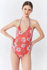 Traje de baño entero con escote en V y tirantes finos, estampado con diseño de flores tipo margarita sobre fondo rojo. Presenta un sistema de tiras cruzadas en la espalda y terminaciones artesanales en resina.