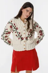 Cárdigan tejido a mano color crudo, con diseño de trenzas y flores bordadas en azul y marrón. Tiene cuello redondo y cierre frontal con botones de madera.