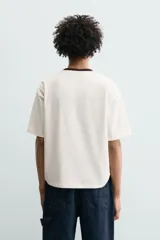 Camiseta de corte relajado confeccionada en algodón, color blanco crudo con cuello redondo en contraste color negro. Presenta un bordado frontal con la frase 'Coração de Melão' y un parche en forma de corazón.