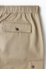 Pantalón cargo de corte holgado en color beige, confeccionado en tejido de algodón. Presenta cintura elástica con cierre de botón a presión, bolsillos laterales al bies, bolsillos de fuelle con solapa en las piernas y pliegues decorativos en las rodillas.