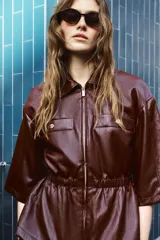 Chaqueta de efecto piel en color bordó, con cuello solapa, cierre frontal con cremallera y mangas por debajo del codo. Presenta cintura elástica y falsos bolsillos delanteros con botones metálicos.