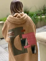Campera tipo hoodie confeccionada en tejido de corderito color camel, con diseño de collage en la espalda que combina parches de diferentes texturas, colores y estampados geométricos. Presenta capucha y corte holgado.