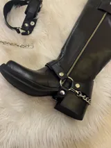Bota alta tipo biker de cuero negro con cierre metálico lateral que recorre toda la caña. Presenta un diseño versátil con estribo y cadena metálica desmontables en la zona del tobillo, taco cuadrado y plataforma delantera.