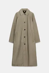 Trench coat largo color verde oliva/beige, con cuello solapa, manga larga ajustable con trabilla y botón, bolsillos delanteros y cierre frontal con botones. El forro interior visible en el cuello es de tela estampada a cuadros en tonos marrones.