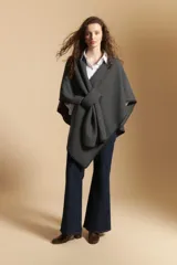 Poncho de tejido suave color beige con diseño envolvente y terminaciones con costuras decorativas en contraste color negro. Presenta un detalle de lazo frontal integrado.