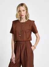 Remera de punto de algodón color terracota, de corte recto, largo estándar y mangas cortas. El diseño incluye superposiciones de tela en los hombros, creando un efecto estructurado y ligeramente geométrico. El cuello redondo presenta ribete acanalado y los laterales tienen pequeñas aberturas para un mejor ajuste.