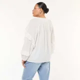 Blusa blanca de manga larga con textura de plumeti (pequeños puntos en relieve) y volantes decorativos verticales en el frente y en los puños. Presenta un escote en V con lazos para anudar en el cuello.