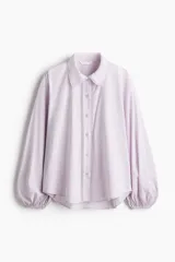Camisa de algodón color lila, con cuello camisero ribeteado en encaje, cierre frontal de botones con detalles de encaje a los lados y mangas globo largas con puños elásticos.