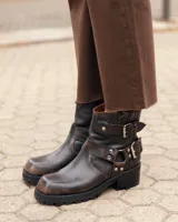Botas de cuero estilo biker en color marrón oscuro con efecto desgastado. Presentan un diseño de caña corta con correas ajustables con hebillas metálicas y detalles de remaches. Cuentan con una suela de goma dentada y tacón bajo.