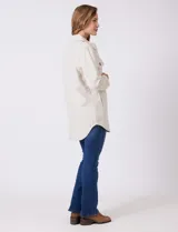 Sobrecamisa de corte amplio confeccionada en tejido suave al tacto, color beige. Presenta cuello de solapa, cierre frontal con botones efecto carey, dos bolsillos frontales con solapa y mangas largas con puños abotonados. El ruedo es redondeado en los laterales y cuenta con canesú en la espalda.