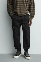Pantalón cargo de corte slim fit en color negro, confeccionado en tejido de algodón con elasticidad. Presenta cintura elástica ajustable con cordón, bolsillos laterales tipo cargo con solapa y bajo acabado en puño elástico.