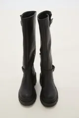 Bota de caña alta color negro, confeccionada en eco cuero, con suela track de goma y detalle de hebilla metálica en el tobillo y en la parte superior de la caña.