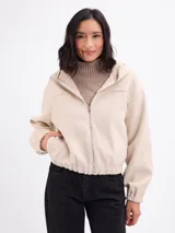 Campera de paño color beige con capucha, cierre frontal completo y mangas largas con puños elásticos. Presenta un diseño corto con ruedo elástico.
