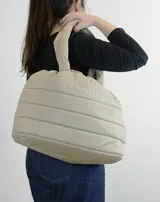 Bolso tipo tote color beige con diseño acolchado y asas tubulares. Presenta el logo de la marca en el centro.
