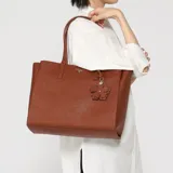 Cartera tipo tote de cuero sintético color marrón de la marca Guess, modelo Lucci. Cuenta con doble asa para el hombro, herrajes dorados, placa triangular con el logo de la marca en el frente y un dije colgante en forma de flor.