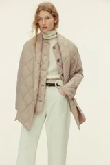 Campera acolchada de color beige con diseño guateado en rombos, hombros caídos, cuello redondo, cierre frontal de botones y bolsillos parche delanteros. Incluye una bufanda a juego que se abrocha en la nuca y cuenta con aislamiento térmico THERMOLITE.