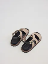 Sandalia de diseño ergonómico con tiras cruzadas en color beige, plantilla negra y suela dentada con pespunte en contraste. Cuenta con una tira trasera ajustable con hebilla metálica.