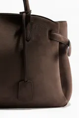 Cartera tote de material tipo gamuza o nobuck, color marrón oscuro, con asas de hombro largas y estructura suave. Presenta tiras decorativas ajustables con remaches en los laterales y una etiqueta colgante extraíble.
