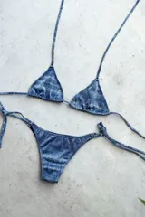 Bikini de dos piezas con estampado azul metalizado o efecto denim desgastado. El corpiño es de estilo triángulo con tiras finas ajustables y apliques de argollas metálicas. La bombacha es vedetina, también de atar en los laterales con apliques de argollas.