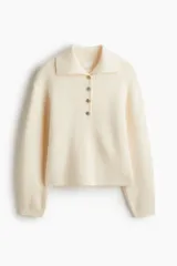 Buzo de punto acanalado color beige, con cuello polo y tapeta frontal con botones metálicos decorativos. Presenta hombros caídos, mangas largas con puños ajustados y diseño de corte recto.