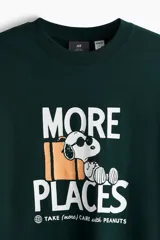 Remera verde oscuro con estampado frontal de Snoopy, equipaje y la inscripción "More Places - Take (more) care with Peanuts". Cuenta con cuello redondo y mangas cortas con hombros caídos.