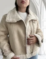 Chaqueta color beige con interior de piel sintética blanca, cierre cruzado con cremallera y detalles de hebillas en los puños.